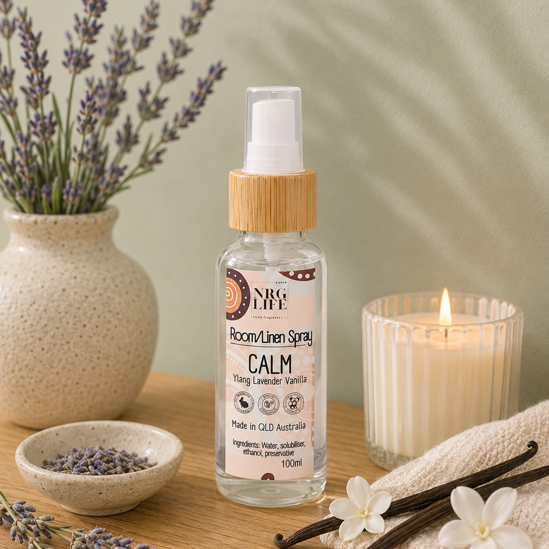 A bottle of Nrglife Calm Room/Linen Spray, styled on a wooden table with lavender, a lit candle, and a cream towel against a soft green background.