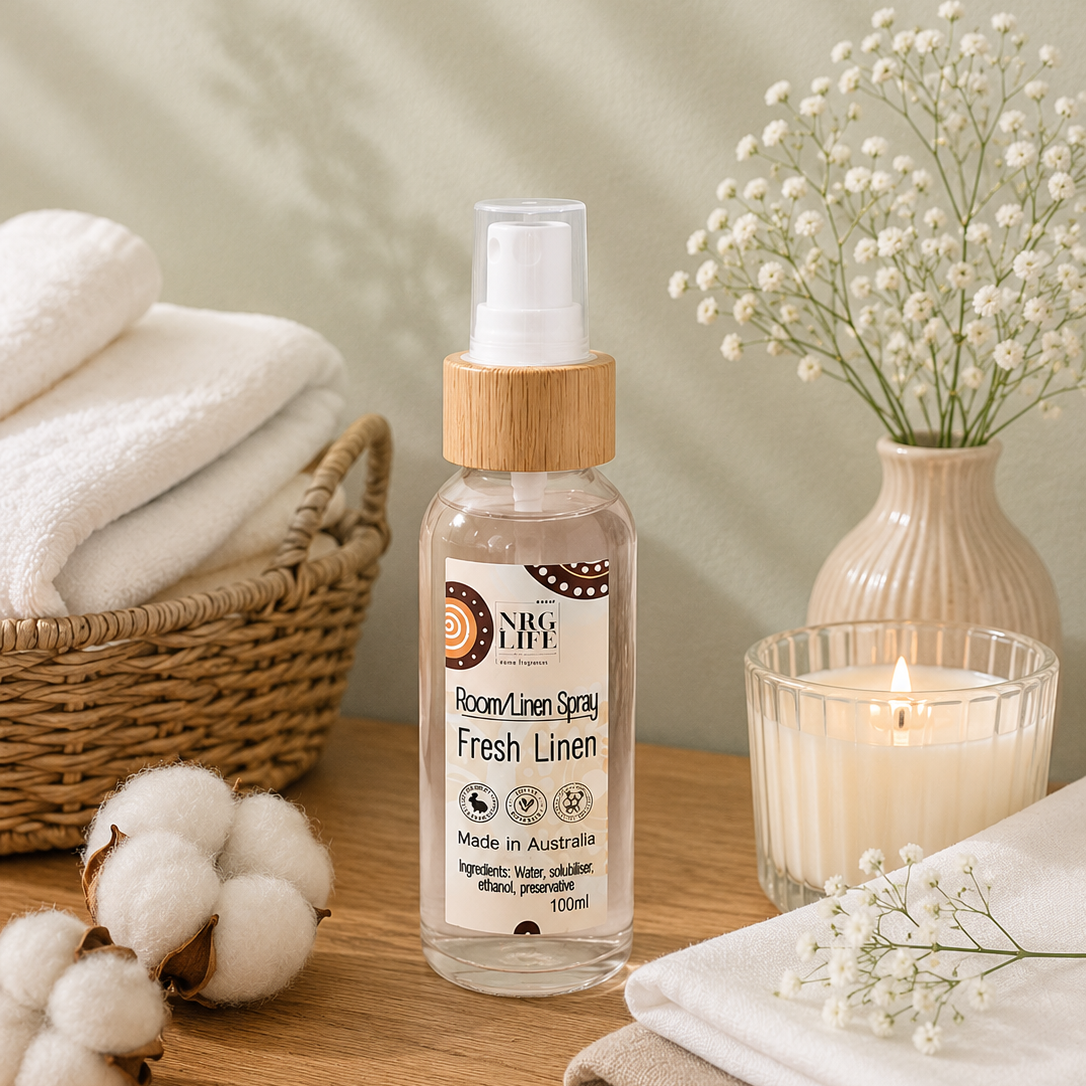 A bottle of Nrglife Room/Linen Spray Fresh Linen placed on a wooden table with folded white towels, cotton bolls, a woven basket, a candle, and a vase of baby's breath flowers.