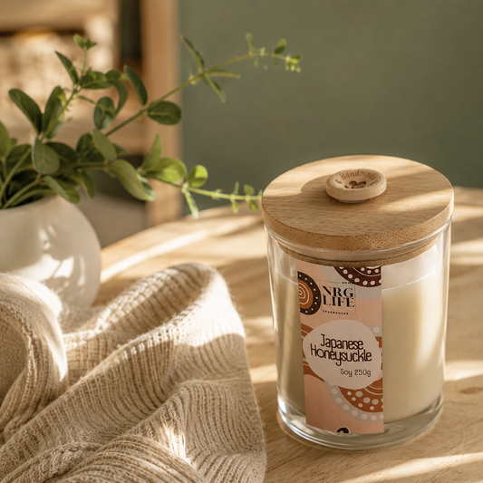 Nrglife's every day candle called Japanese Honeysuckle. It's a glass jar with a timber top and an Australian Label sitting on a timber table with greenery filtered light and a green wall background.