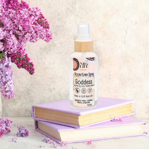A room linen spray sitting on a hallway table on top of two books with a bunch of flowers to the side. The table and backdrop wall all in neutral beige shades.