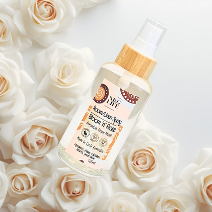 Bottle of 'Bloom 'n Rose' room/linen spray with a wooden cap on a bed of white roses.