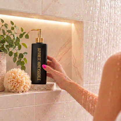 500ml Triangle Black Coconut Body Wash bottle with a fragrance called Japanese Honeysuckle, sitting in a shower niche with a loofah beside it, the shower is running and a female's hand is reaching out to grab it.