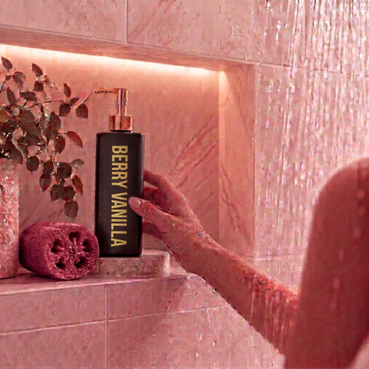 A hand holding a black pump body wash bottle 500ml labeled 'BERRY VANILLA' with gold lettering, set against a pink marble shower niche with a pink sponge and dried leaves.