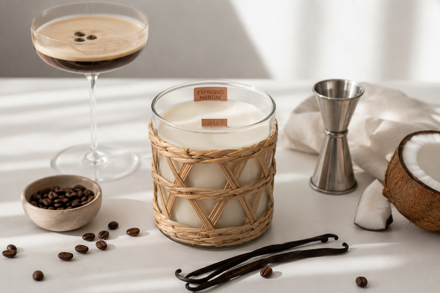 Candle with rattan weave overlay, glass of coffee, coconut, and vanilla beans on a light surface.