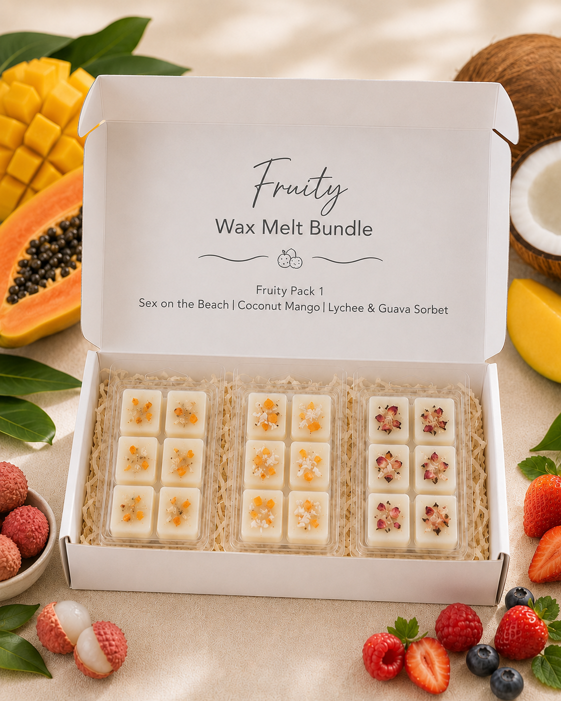 An open white gift box labeled 'Fruity Wax Melt Bundle' containing three rows of small white wax melts with citrus and floral inclusions, styled with sliced mango, papaya, strawberries, raspberries, blueberries, and coconut on a beige fabric background.
