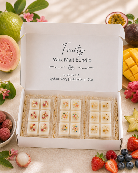 An open white gift box labeled 'Fruity Wax Melt Bundle' containing three trays of small white wax melts decorated with dried flower petals and colorful sprinkles. The box is surrounded by fruit slices and berries on a light beige surface.