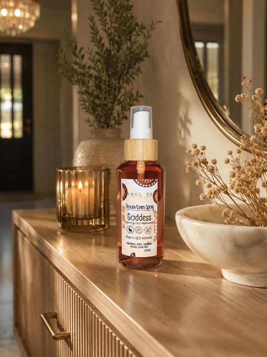 A bottle of 'Goddess' room linen spray with amber liquid and a bamboo pump, displayed on a wooden console table in a warm, decorated interior setting.