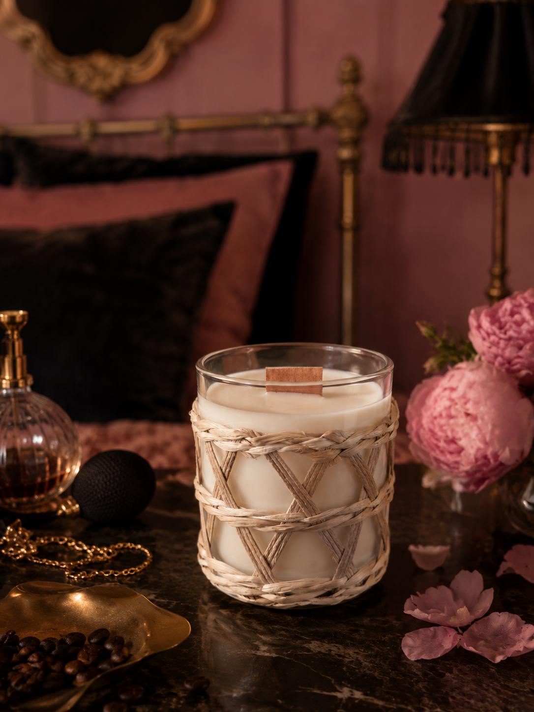 A glass container wrapped in woven rattan. Placed on a black marble table with flowers in the background, perfume set in a bedroom with a four poster bed, pink walls and golden mirror and accessories.