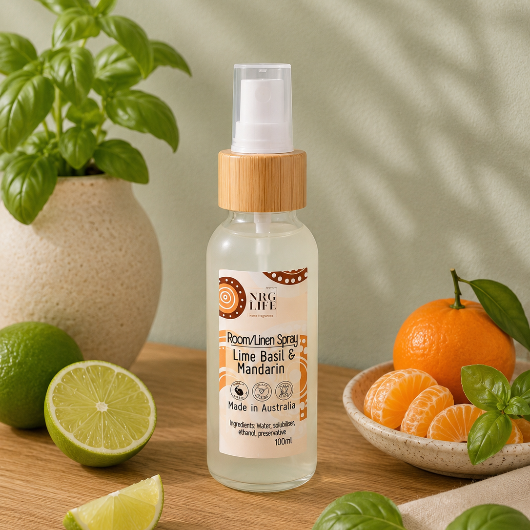 A 100ml room linen spray bottle by Nrglife, scented with Lime, Basil, and Mandarin (Essential Oils), inspired by Jo Malone, displayed on a wooden table surrounded by fresh limes, oranges, and basil leaves against a soft green leaf-shadowed background.