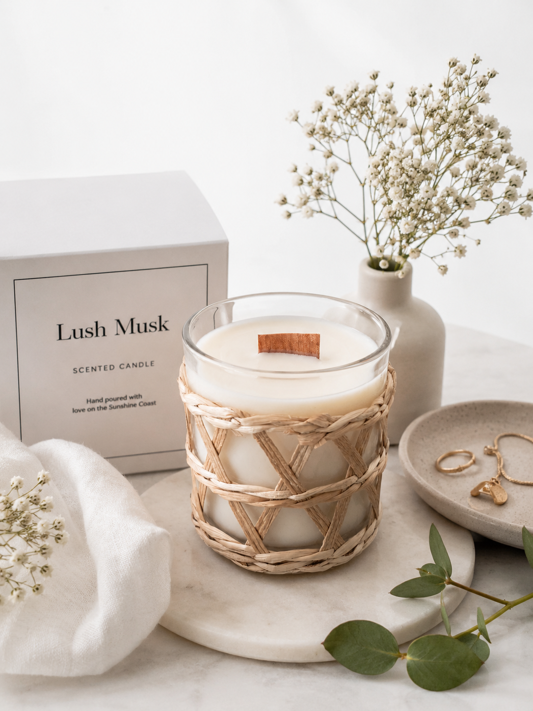 Candle with timber wick, called 'Lush Musk'. A clear glass container surrounded by a rattan cane overlay and decorative elements on a marble surface.