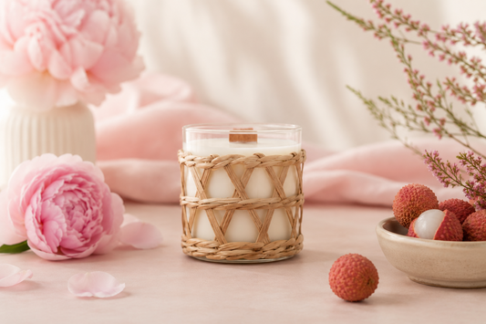 Coconut Soy Candle with the scent Lychee & Peony in a glass jar with a rattan weave overlay. Placed on a table with pink flowers and lychees on a soft feminine background.