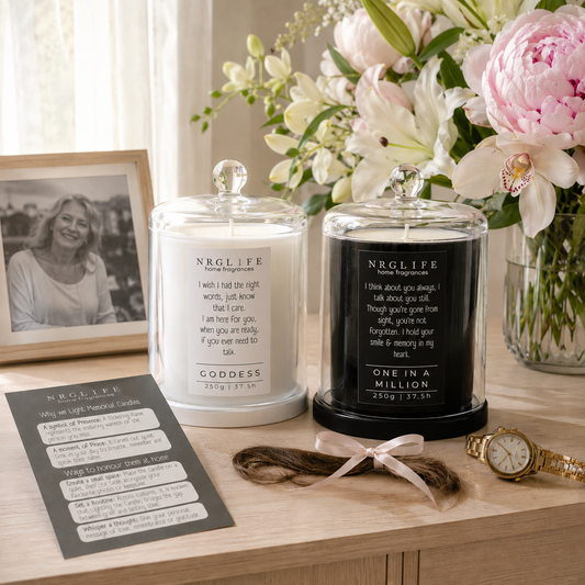 Two NGRLIFE memorial candles in glass jars with lids, displayed on a wooden table alongside a hair sample, a watch, a framed photo, and a card. One candle is white with a clear glass cloche cover with a Goddess scent and the other is black with a clear glass cloche cover with One Million scent.