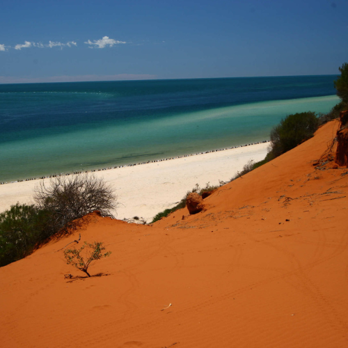 Red sand dune leading to a beach with clear blue water and sky.