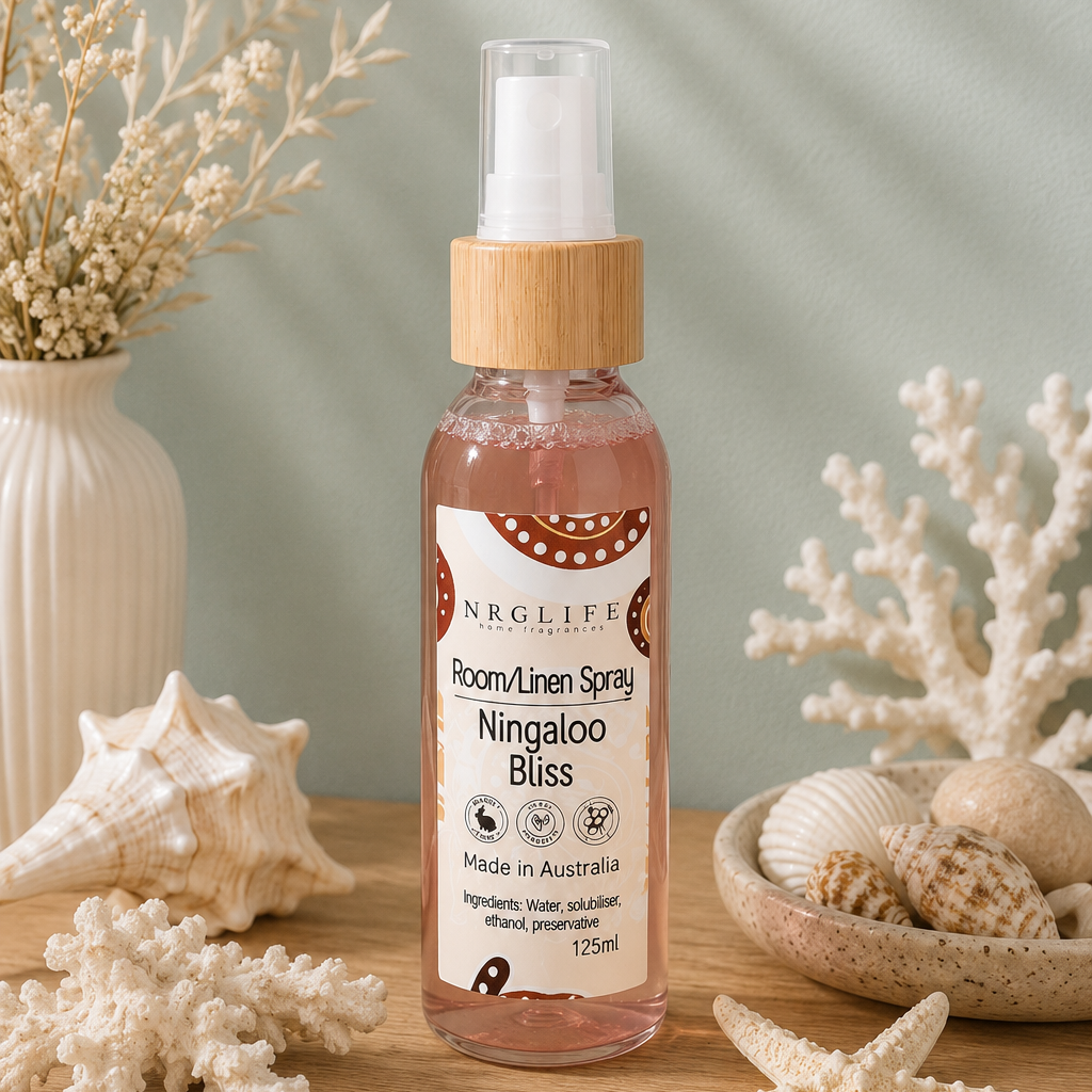 A bottle of Nrglife Room/Linen Spray, specifically 'Ningaloo Bliss', styled on a wooden surface with seashells, starfish, dried flowers, and a vase against a soft green background with leaf shadows.