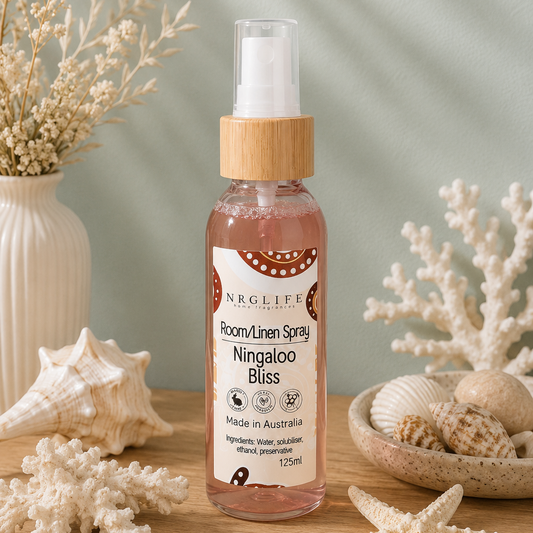 A bottle of Nrglife Room/Linen Spray, specifically 'Ningaloo Bliss', styled on a wooden surface with seashells, starfish, dried flowers, and a vase against a soft green background with leaf shadows.