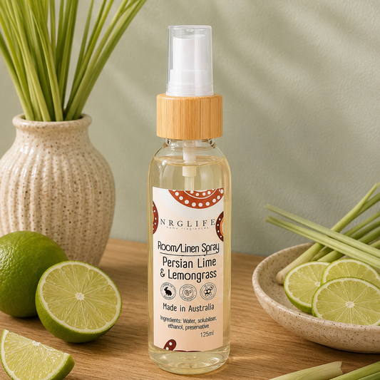 A bottle of Nrglife Room/Linen Spray with Persian Lime and Lemongrass scent, styled on a wooden table with limes, lemongrass stalks, and a potted plant.