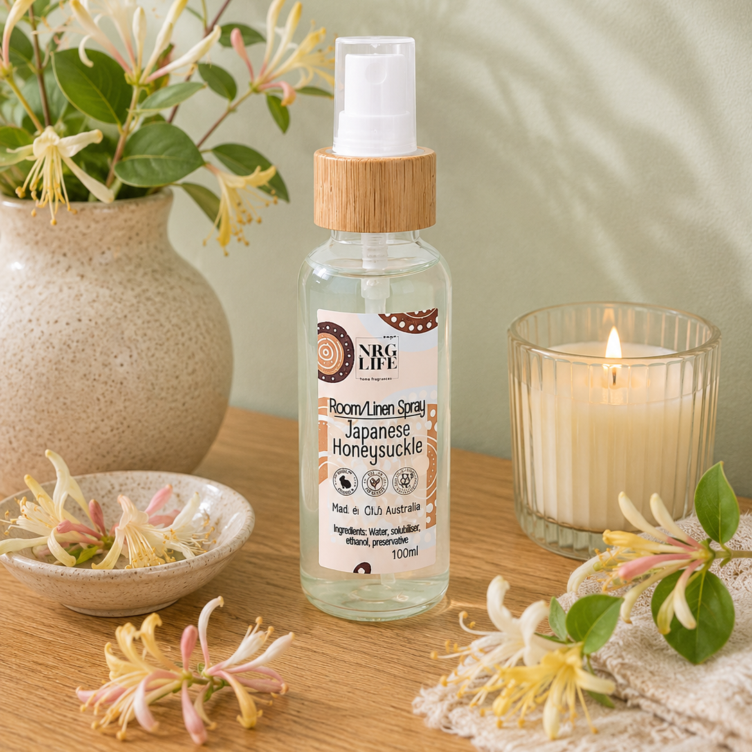 A 120ml plastic bottle of Nrglife Japanese Honeysuckle Room Linen Spray sits on a wooden table, styled with a vase of hibiscus flowers, a lit candle in a glass holder, and scattered flower petals.