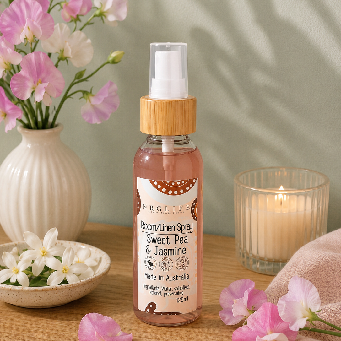 A bottle of Nrglife Room Linen Spray (Sweet Pea & Jasmine) styled on a wooden table with pink flowers, a white vase, a lit candle, and a soft pink cloth.